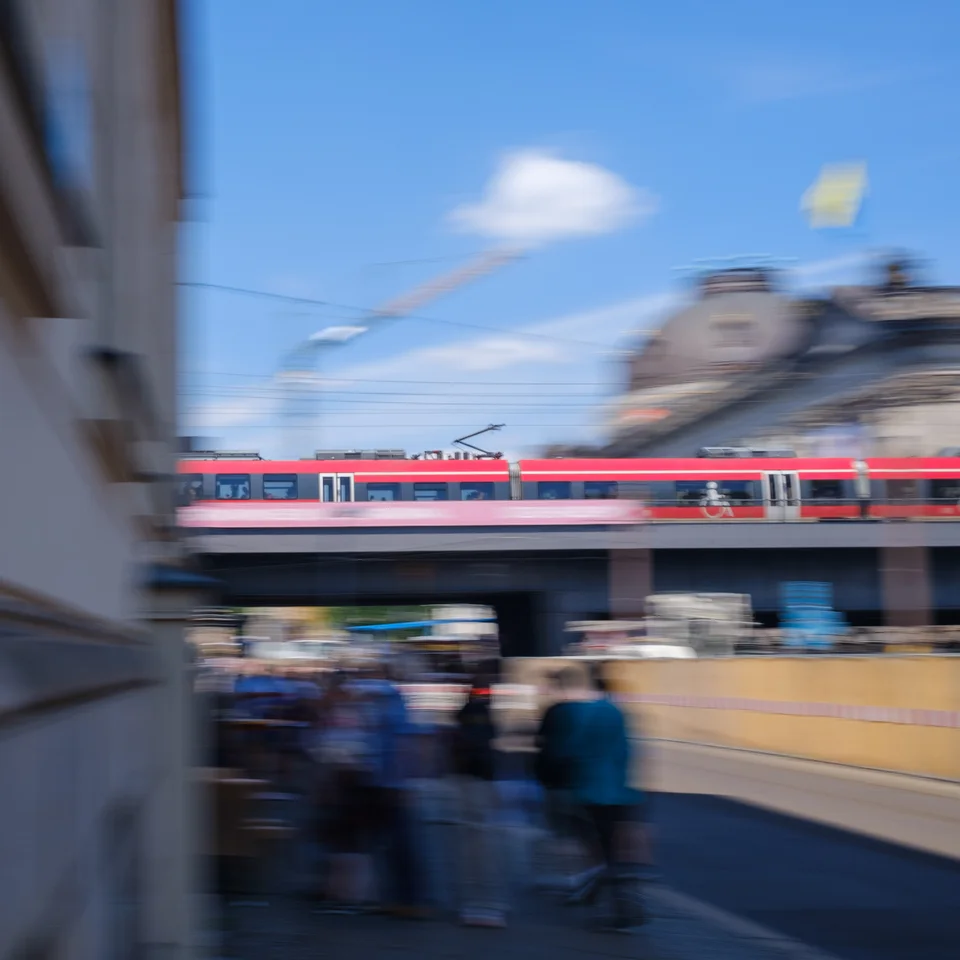 A red train crossing above a busy Berlin street, with motion blur around the buildings and pedestrians.